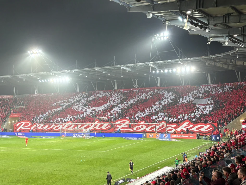 Oprawa kibiców Widzewa Tłum kibiców na stadionie tworzy efektowną choreografię w biało-czerwonych barwach, trzymając kartoniki z napisem 'Rewolucja Łódzka', jasne oświetlenie, rozgrywka piłkarska na boisku.