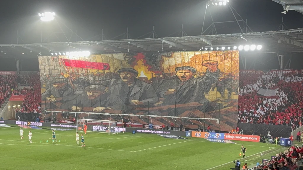 Oprawa kibiców Widzewa Ogromna sektorówka z historycznym propagandowym motywem przedstawiająca postacie stylizowane na bolszewików, trzymana przez kibiców na stadionie piłkarskim podczas meczu; w tle widoczne trybuny wypełnione czerwono-białymi flagami.