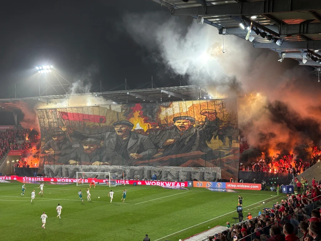 Łódź - miasto buntu Tłum kibiców na stadionie piłkarskim tworzy monumentalną oprawę z historycznym motywem i polską flagą, w tle rozbłyskują race i kłębi dym, a na murawie toczy się mecz.