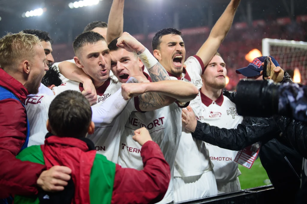 Grupa piłkarzy w białych strojach świętuje zwycięstwo na stadionie, wyrażając silne emocje, z uniesionymi ramionami, okrzykami i gestami radości, otoczona kibicami przebranymi w klubowe barwy.