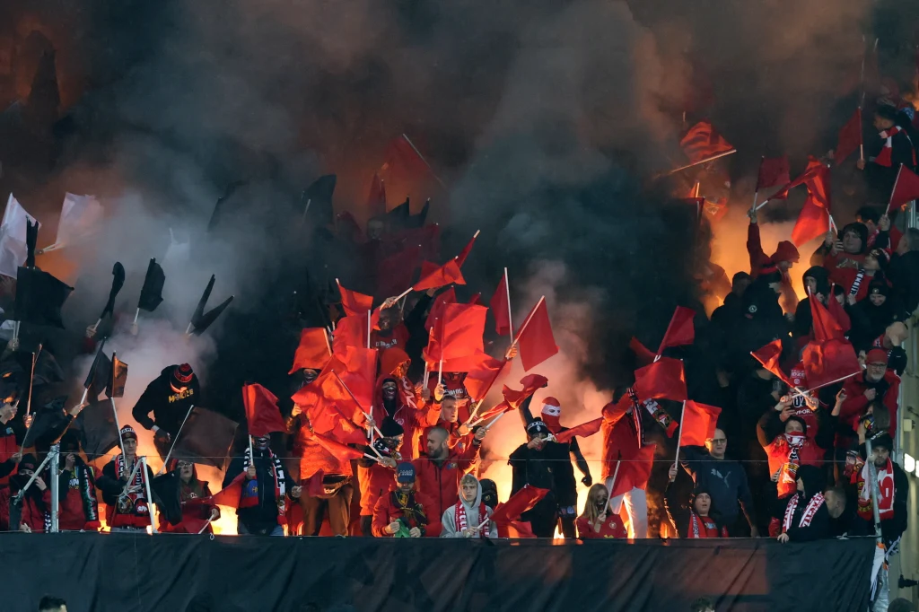 Tłum kibiców w czerwonych barwach macha flagami, a w tle unoszą się kłęby dymu z odpalonych rac, rozświetlając przestrzeń intensywnym pomarańczowym światłem.