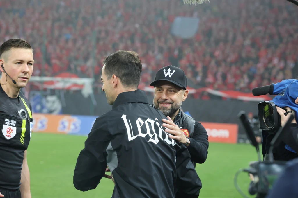 Sędzia piłkarski, trener drużyny oraz inna osoba wymieniają uściski i gesty powitania na stadionie podczas meczu piłkarskiego, w tle widoczna publiczność na trybunach.