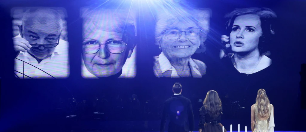 Trzy osoby stoją na scenie i patrzą na duży ekran z czterema portretami starszych ludzi, silne reflektory oświetlają przestrzeń tworząc uroczysty nastrój.