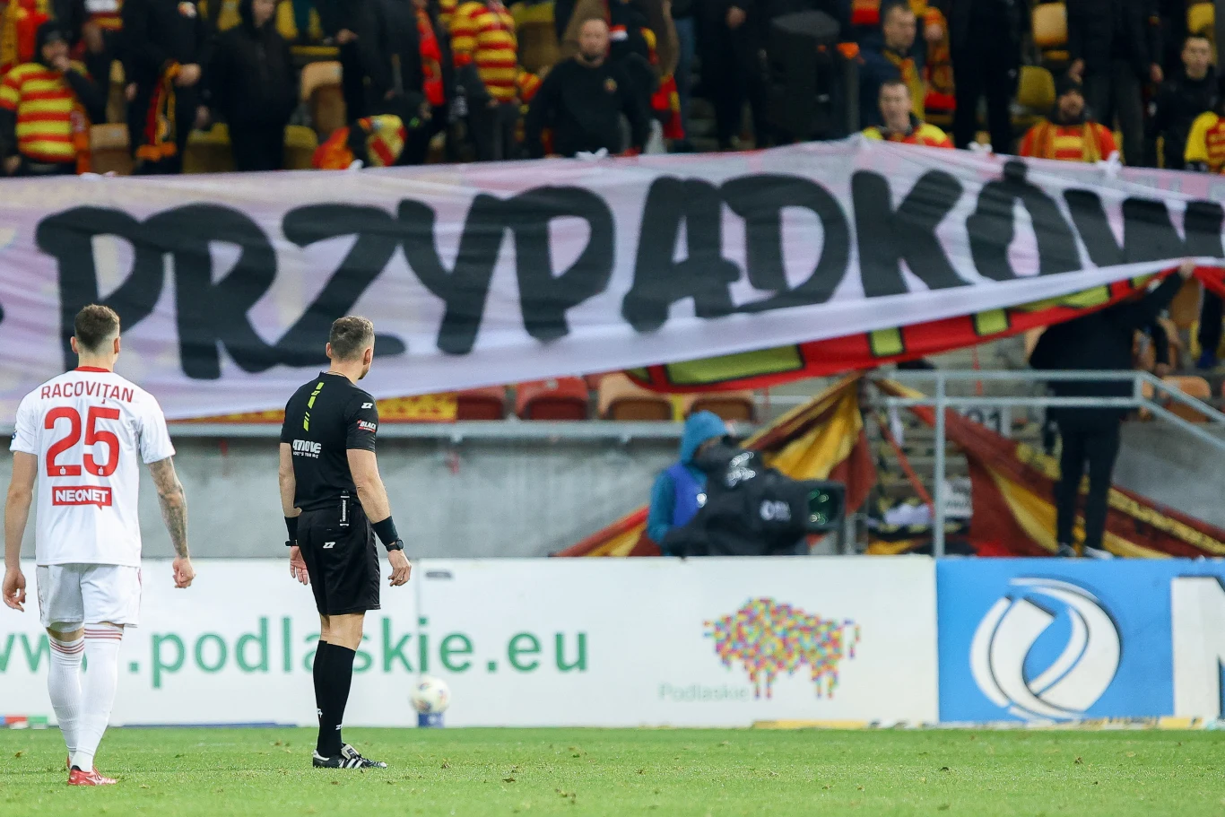 Paweł Raczkowski przerwał spotkanie Jagiellonia Białystok - Raków Częstochowa z powodu transparentu obrażającego innego z arbitrów Fragment meczu piłkarskiego, na którym znajduje się zawodnik w białym stroju z numerem 25, arbiter oraz widoczny w tle duży transparent z napisem PRZYPADKOWI, trzymany przez kibiców na trybunach stadionu.