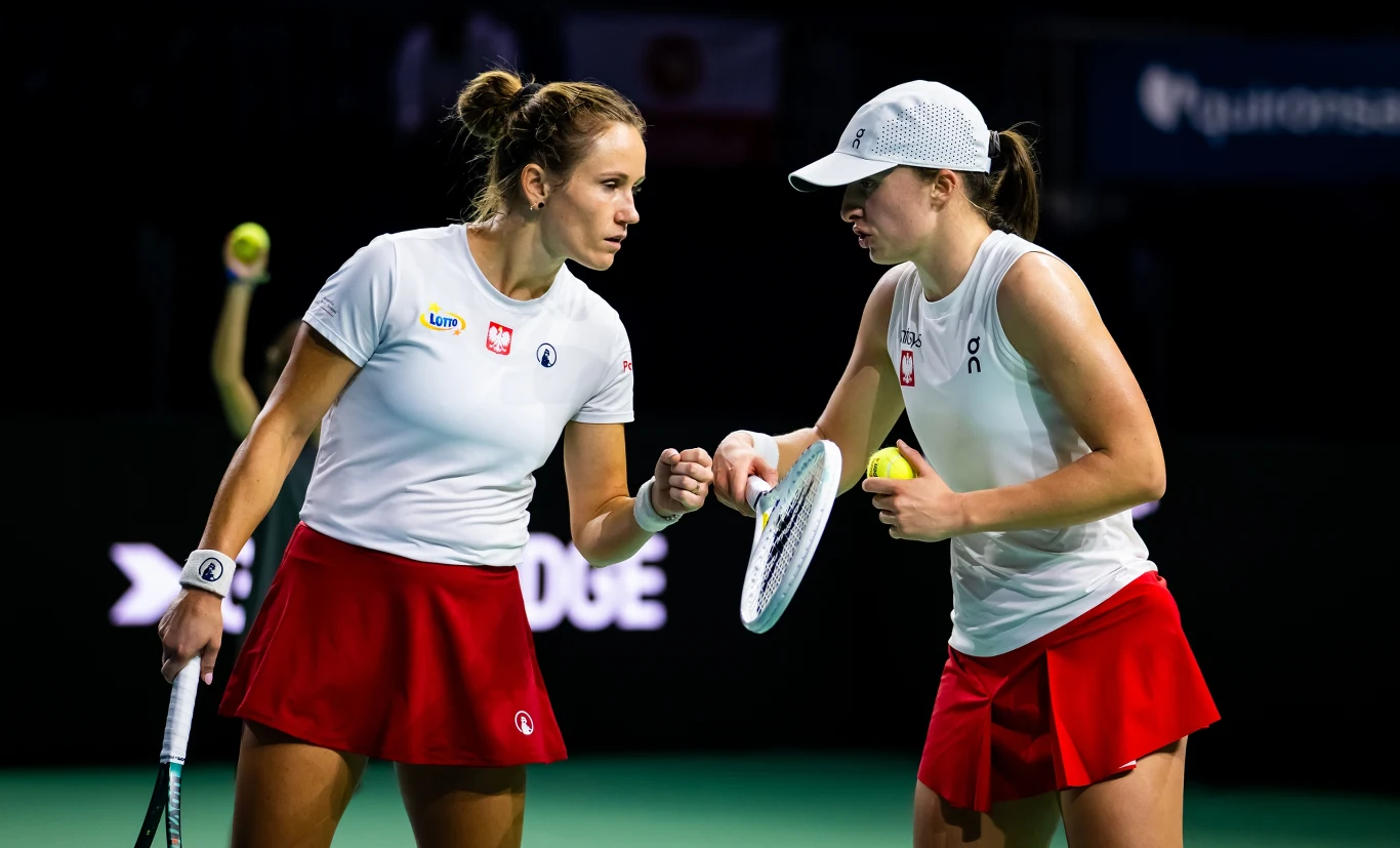 Katarzyna Kawa awansowała w rankingu WTA o jedną pozycję, Iga Świątek wciąż pozostaje druga. Za półtora tygodnia obie pojawią się w Gorzowie Dwie tenisistki w białych koszulkach i czerwonych spódniczkach stoją na korcie, komunikując się ze sobą podczas meczu deblowego. Jedna z nich trzyma rakietę i piłkę tenisową, obie są skupione na grze.