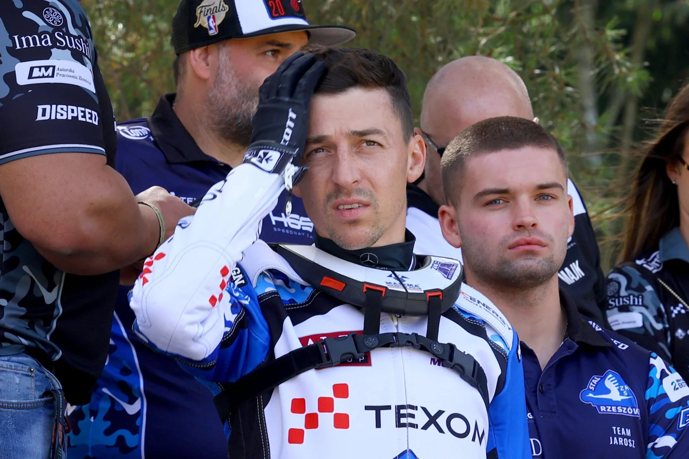 Kierowca w stroju motocrossowym marki Texom trzyma rękę na głowie, otoczony przez kilku mężczyzn ubranych w koszulki zespołowe. Na twarzach widać skupienie i lekkie napięcie, w tle pojawia się zieleń.