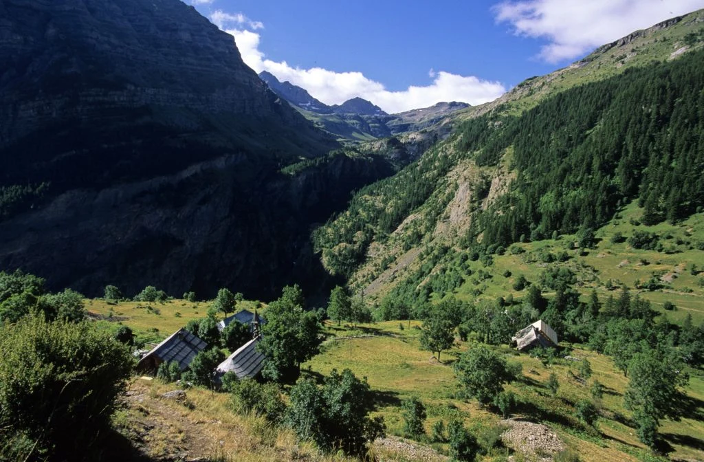 Zielona dolina górska z rozproszonymi domkami, otoczona gęstym lasem i stromymi zboczami. W oddali widoczne majestatyczne, skaliste szczyty, a krajobraz oświetlony jest jasnym światłem dziennym.