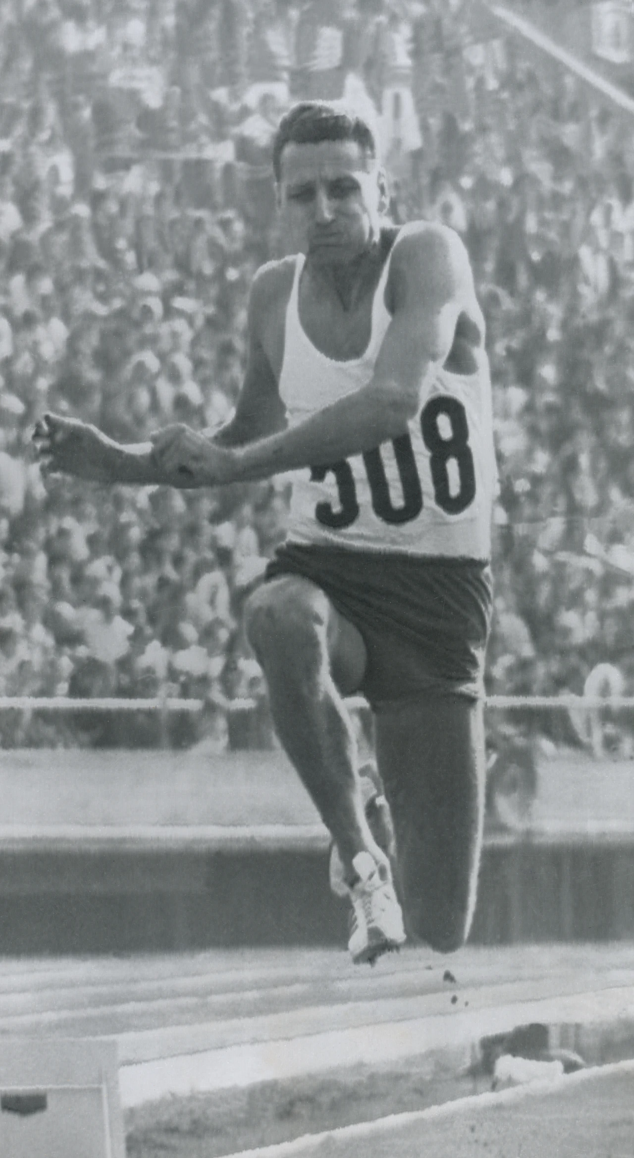 Lekkoatleta ubrany w biały podkoszulek i ciemne spodenki, z numerem startowym 308, wykonuje skok w dal na stadionie lekkoatletycznym w obecności licznej publiczności.