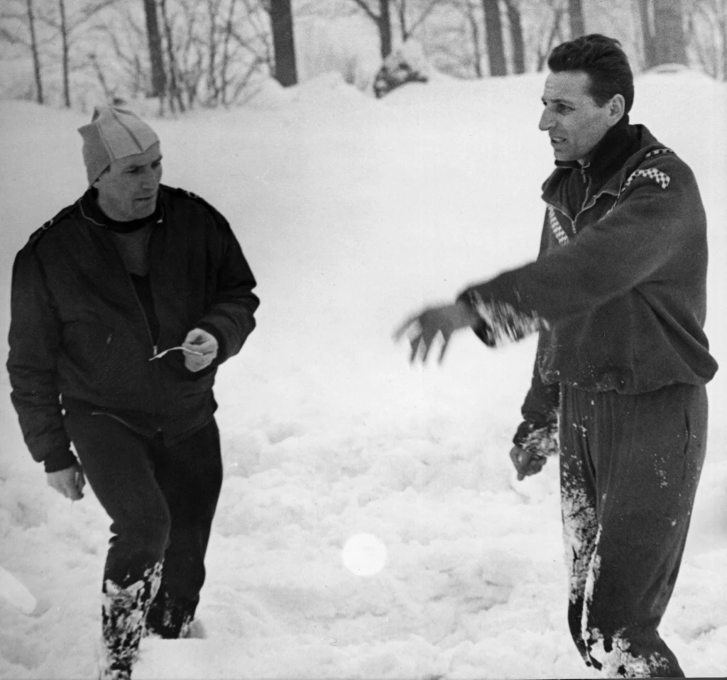 Dwóch mężczyzn w sportowych ubraniach stoi w głębokim śniegu, w tle widoczne są drzewa pokryte śniegiem, jeden z nich wykonuje gest ręką, drugi trzyma drobny przedmiot