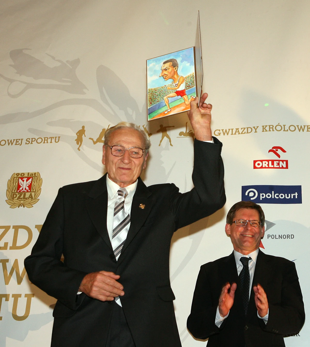 Starszy mężczyzna w garniturze podnosi w górę rysunek przedstawiający sportowca, obok niego stoi mężczyzna w okularach, również elegancko ubrany, bijący brawo. Za nimi widoczne są logotypy sponsorów i złote napisy związane ze sportem.