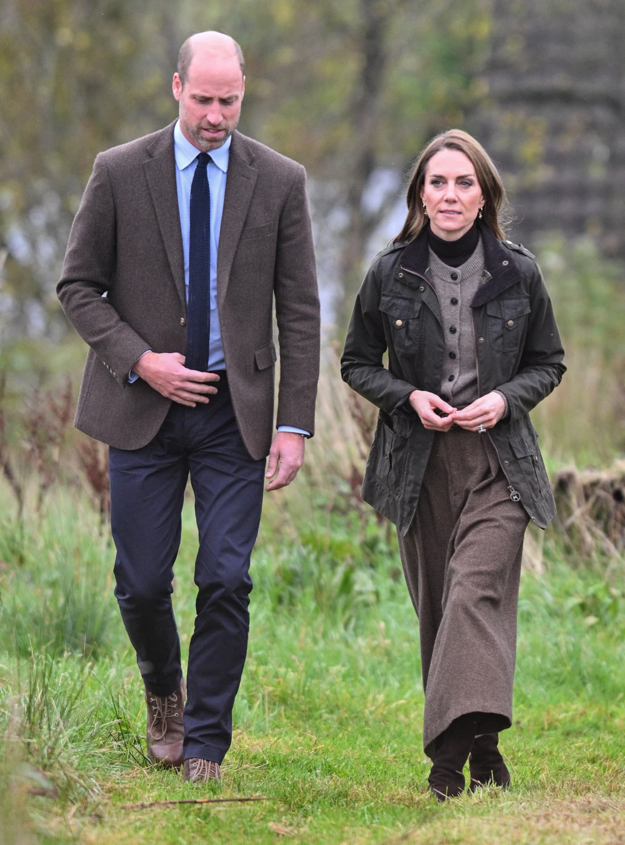 Para ubrana w eleganckie jesienne stroje spaceruje po zielonej trawie, otoczona naturalnym krajobrazem z rozmytym tłem roślinności.