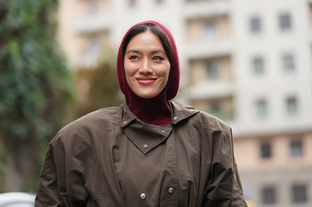 Kobieta o ciemnych włosach uśmiecha się lekko, ubrana w bordowy golf i brązową kurtkę z szerokimi ramionami, w tle widać rozmyte budynki.