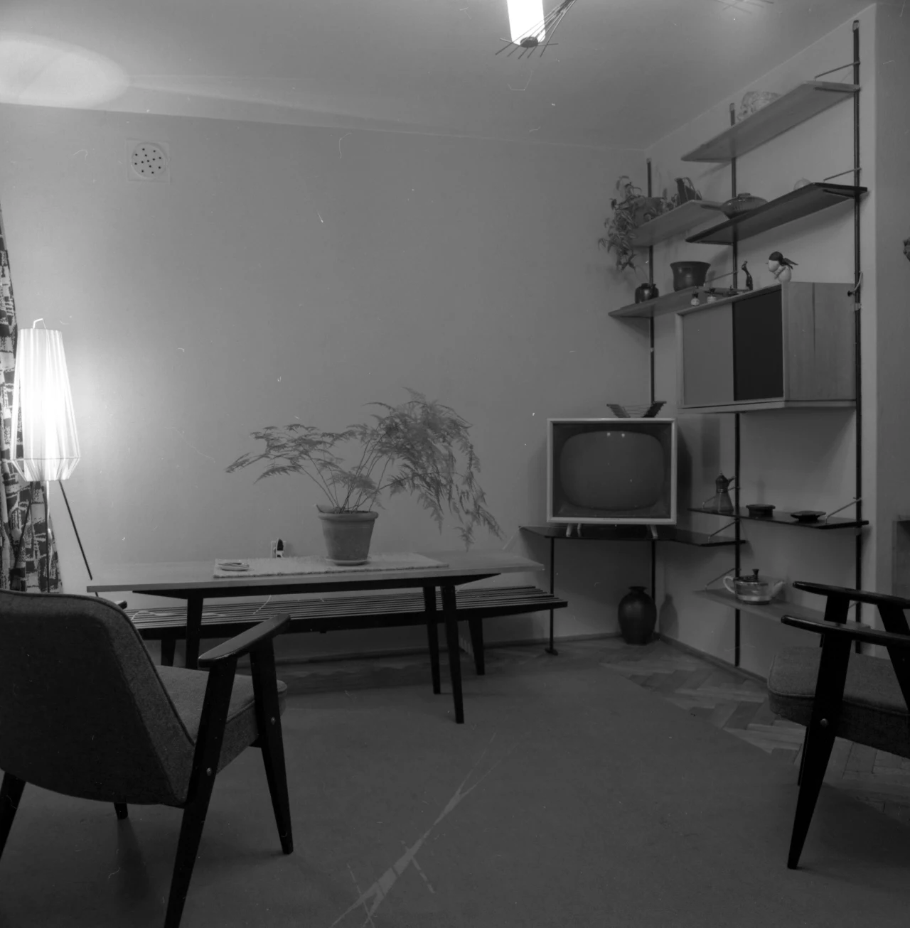 Ciemny, minimalistycznie urządzony salon z dwoma fotelami, ławą, starą telewizją kineskopową na regale oraz dużą lampą podłogową w rogu, na stole doniczka z rośliną, wnętrze w stylu lat 60.