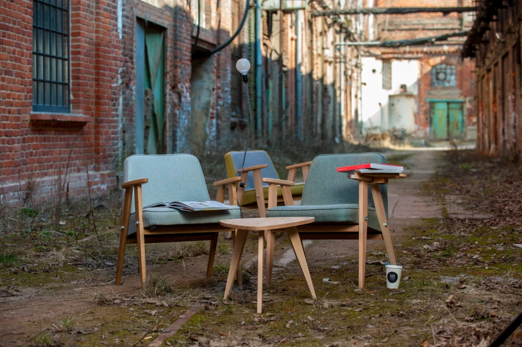 Dwa fotele i stolik kawowy ustawione w opuszczonym, zrujnowanym budynku z czerwonej cegły, wokół widać ślady zniszczenia oraz porastającą ziemię roślinność, a na stole leżą czasopisma i kubek jednorazowy.