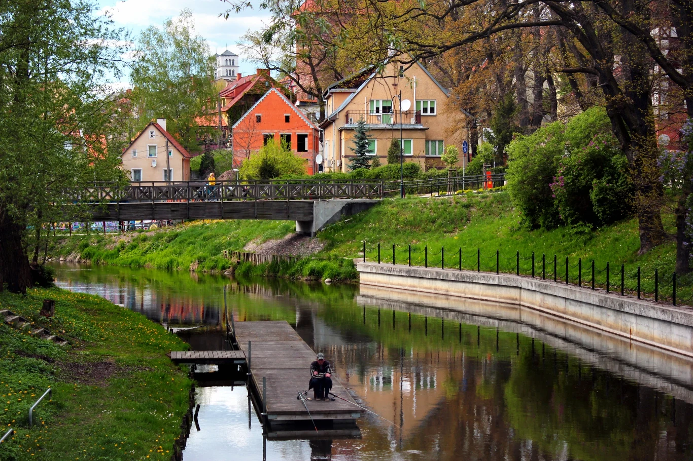 Lidzbark Warmiński to urokliwe i nieodkryte miasto, które zachwyca nie tylko klimatem, ale bogatą historią i wieloma atrakcjami Rzeka otoczona zielenią i drzewami, z drewnianym pomostem, na którym siedzi osoba. W tle malownicze, kolorowe kamienice oraz most dla pieszych. Spokojna, wiosenna atmosfera miasta.