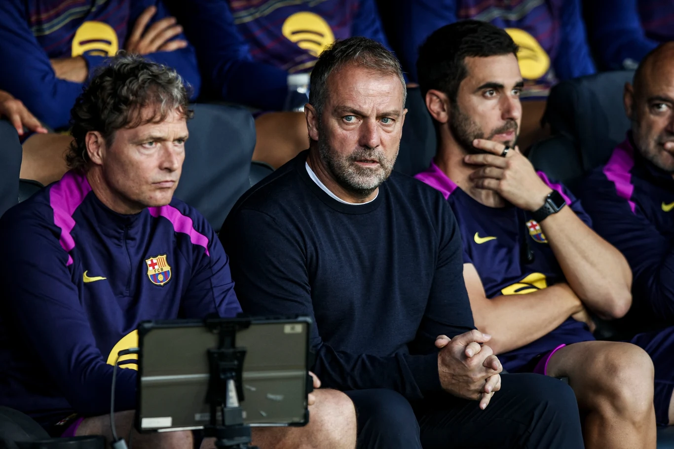 Hansi Flick Grupa trenerów klubu FC Barcelona w oficjalnych strojach siedzi skupiona na ławce, obserwując wydarzenia na boisku. Pierwszy plan zajmuje mężczyzna w czarnym swetrze, po bokach współpracownicy w fioletowych bluzach z logo klubu.
