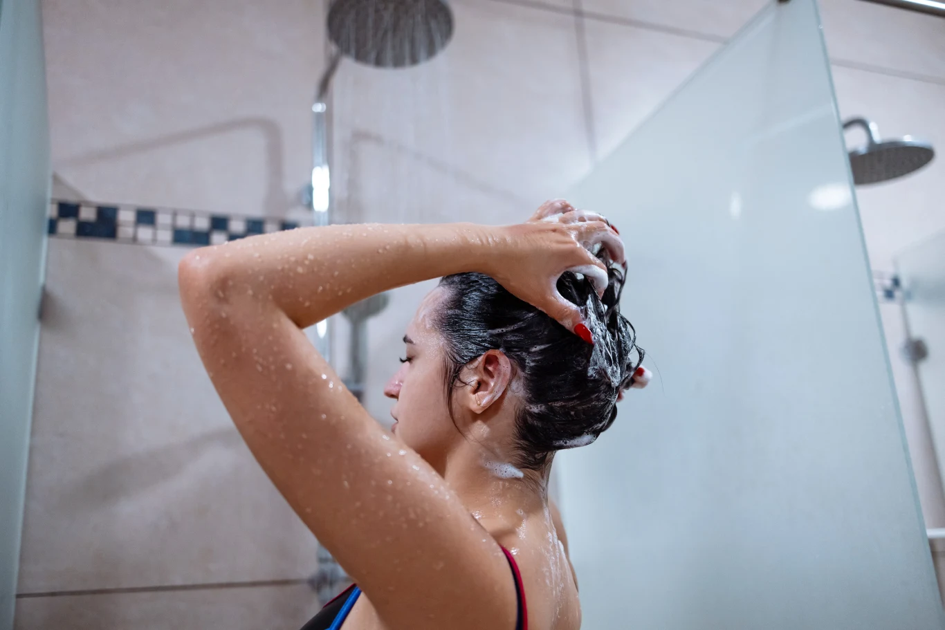 Nie tylko suszarka. Gorąca woda też osłabia włosy i przyspiesza ich wypadanie Kobieta myje włosy pod prysznicem, spływają na nią krople wody, wokół widoczne są szklane ścianki kabiny prysznicowej i płytki ścienne.