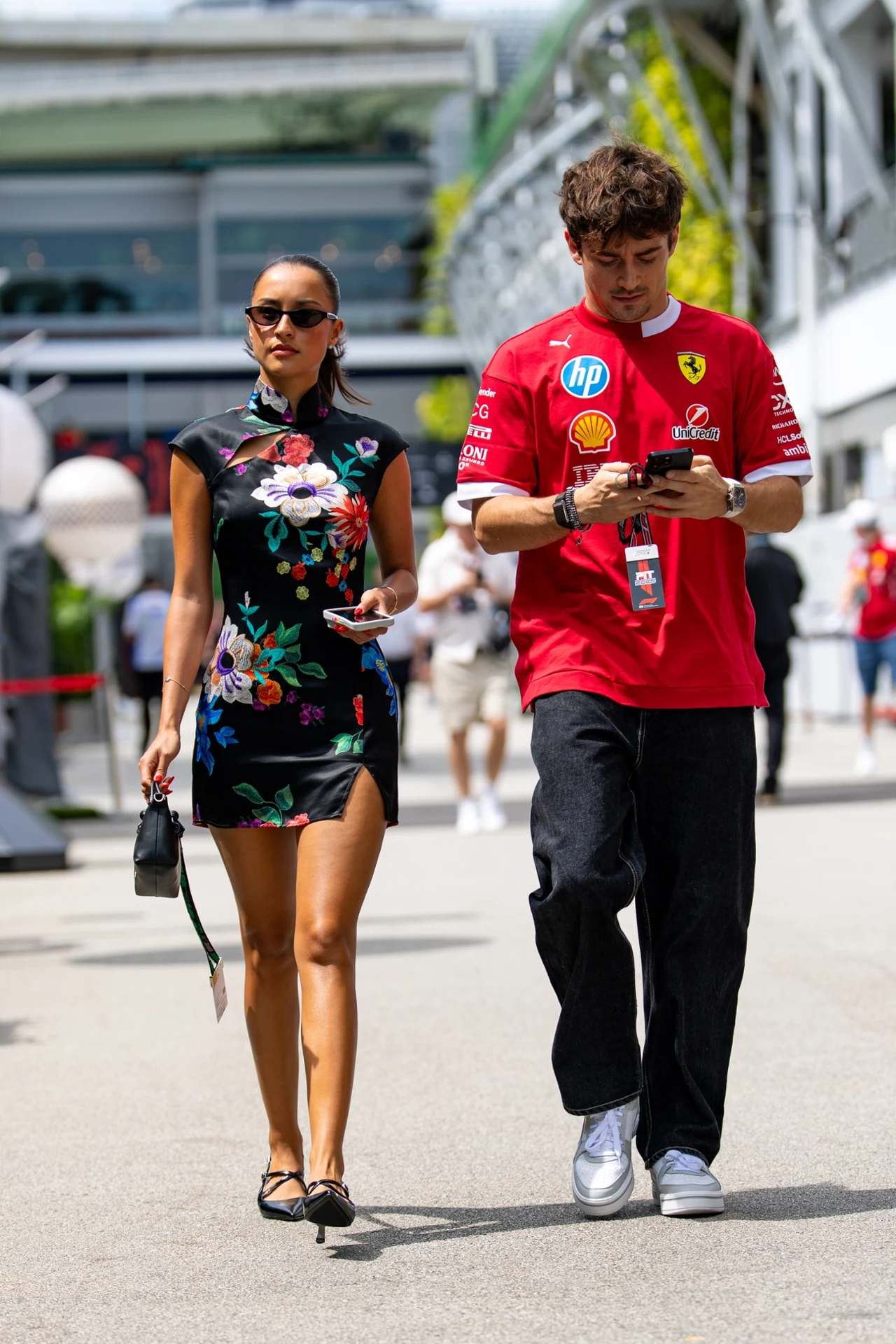 Charles Leclerc i Alexandra Saint Mleux Para spacerująca promenadą, kobieta w eleganckiej, czarnej sukience z kwiatowym wzorem i okularach przeciwsłonecznych, mężczyzna w czerwonej koszulce Ferrari, skupiony na telefonie. W tle widać obiekt sportowy oraz innych ludzi.