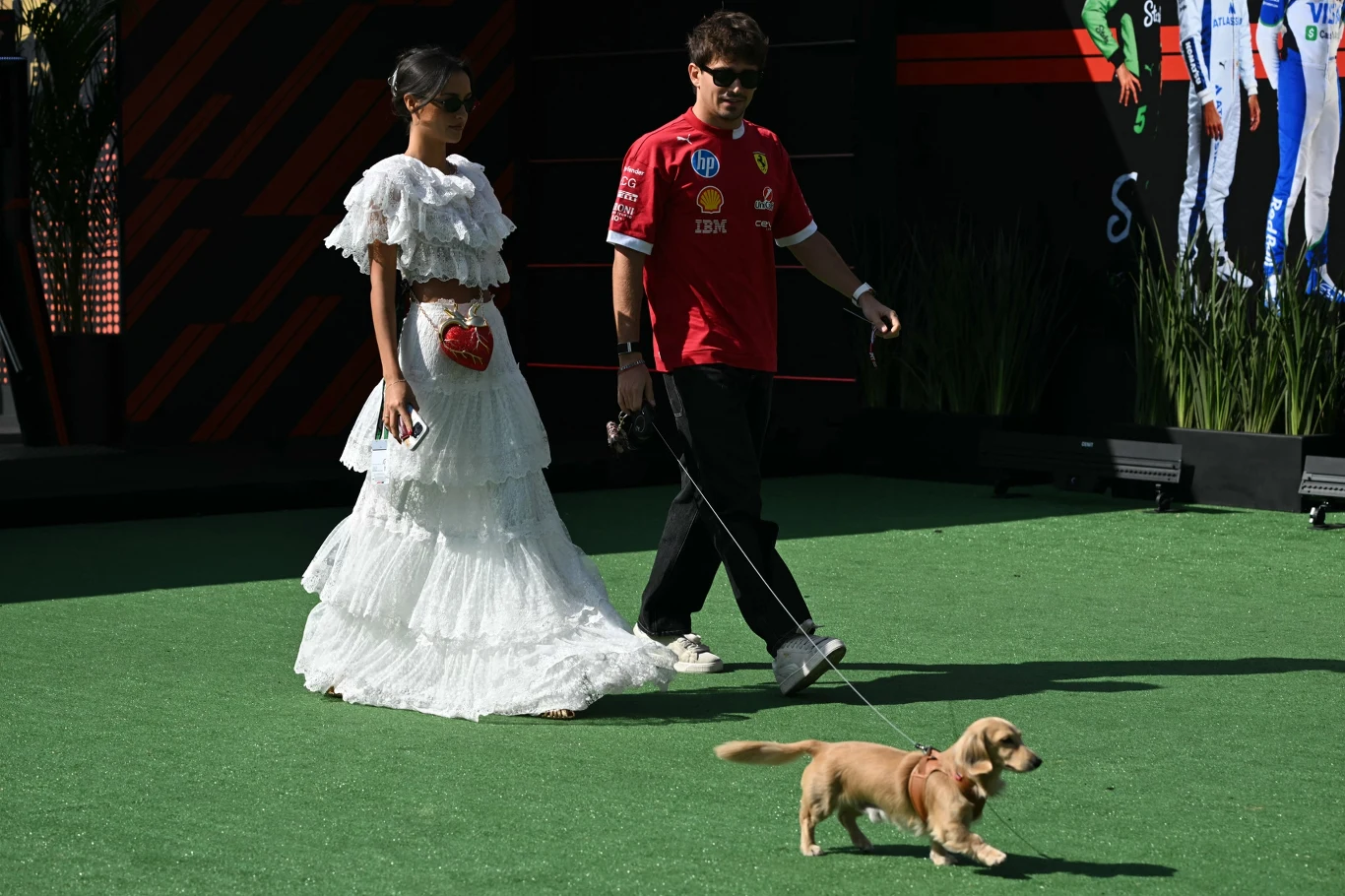 Charles Leclerc i Alexandra Saint Mleux Kobieta w białej sukni z czerwonym sercem na biodrze oraz mężczyzna w czerwonej koszulce prowadzą na smyczy małego psa po zielonym trawniku. W tle widoczne ciemne elementy dekoracyjne oraz fragmenty postaci na grafice.