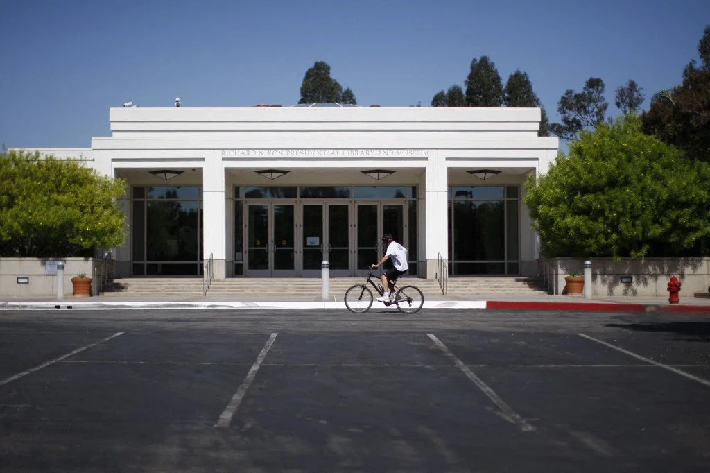 Biały budynek o nowoczesnej architekturze otoczony drzewami, na pierwszym planie pusta nawierzchnia parkingu oraz osoba jadąca na rowerze przed wejściem głównym.