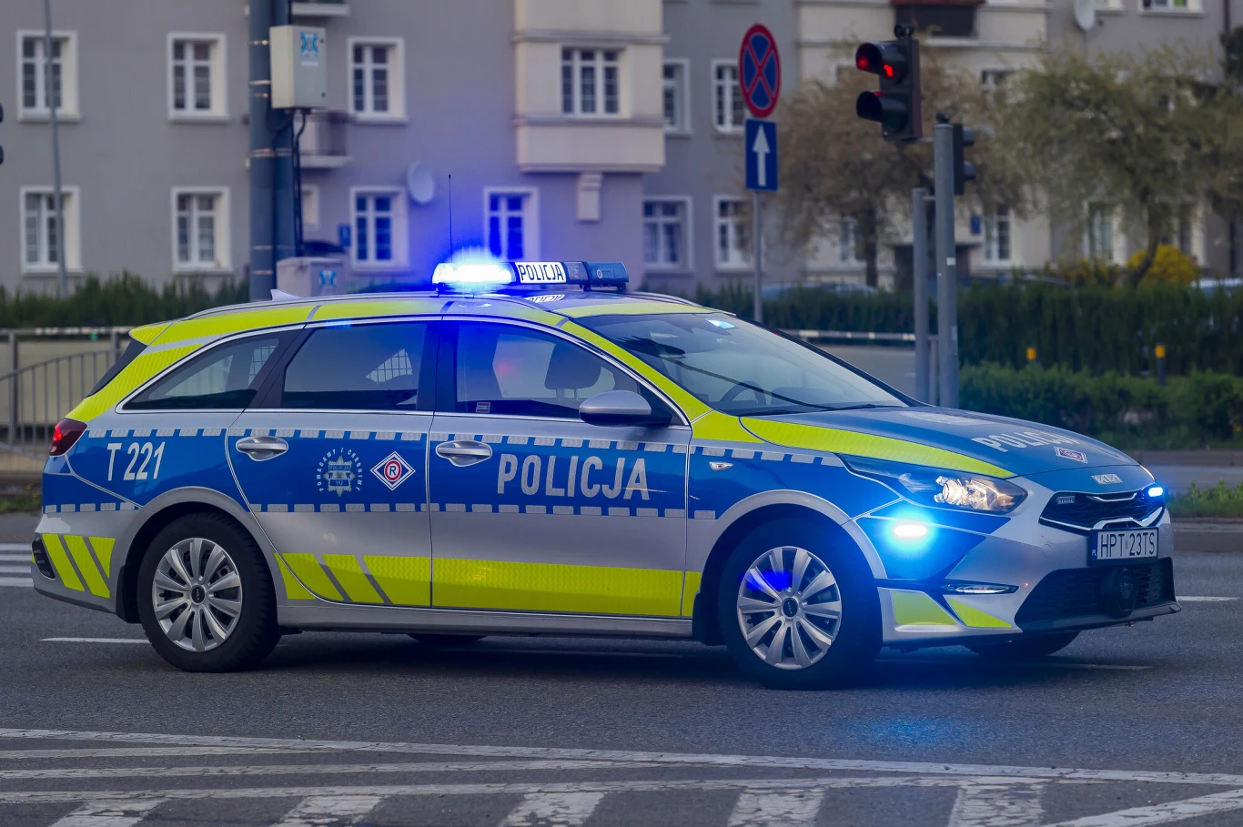 Gdynia. "Nazar" zatrzymany przez policję (zdj. ilustracyjne) Policyjny radiowóz z włączonymi niebieskimi światłami jedzie przez skrzyżowanie w mieście, w tle widoczne są budynki mieszkalne oraz sygnalizacja świetlna.