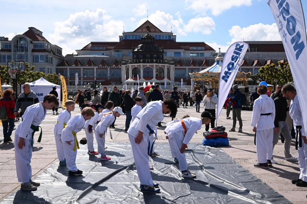 Czyste, Zielone Miasta w Sopocie Grupa dzieci i młodzieży ubranych w białe stroje do karate wykonuje ukłon na macie rozłożonej na rynku, w tle widać budynki, flagi oraz przechodniów obserwujących pokaz.