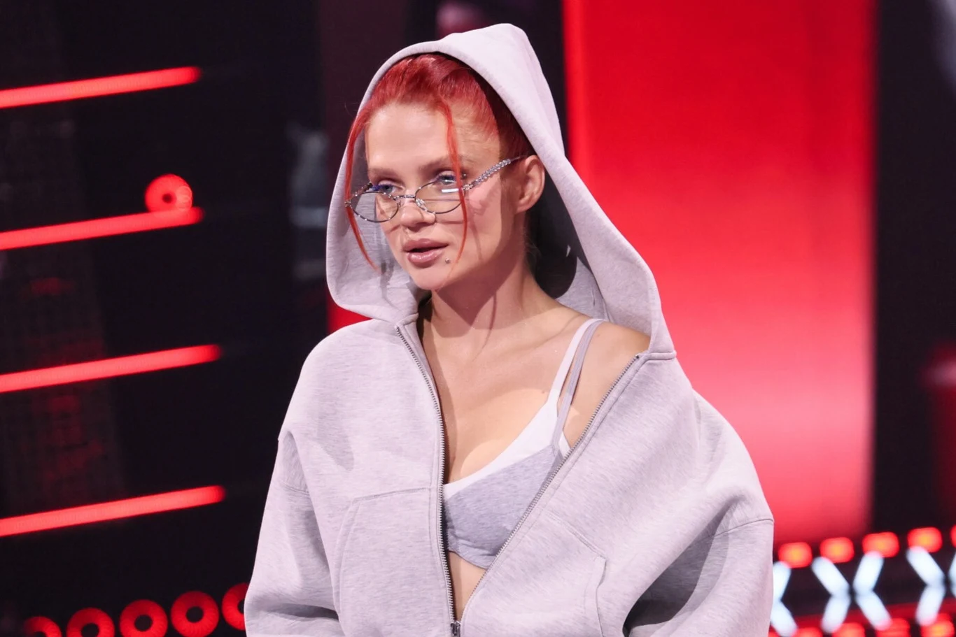 Decyzja Margaret wywołała burzę w sieci Kobieta o rudych włosach w szarej bluzie z kapturem oraz sportowym staniku, nosząca okulary o przeźroczystych oprawkach, stoi na tle czerwono-czarnej sceny.