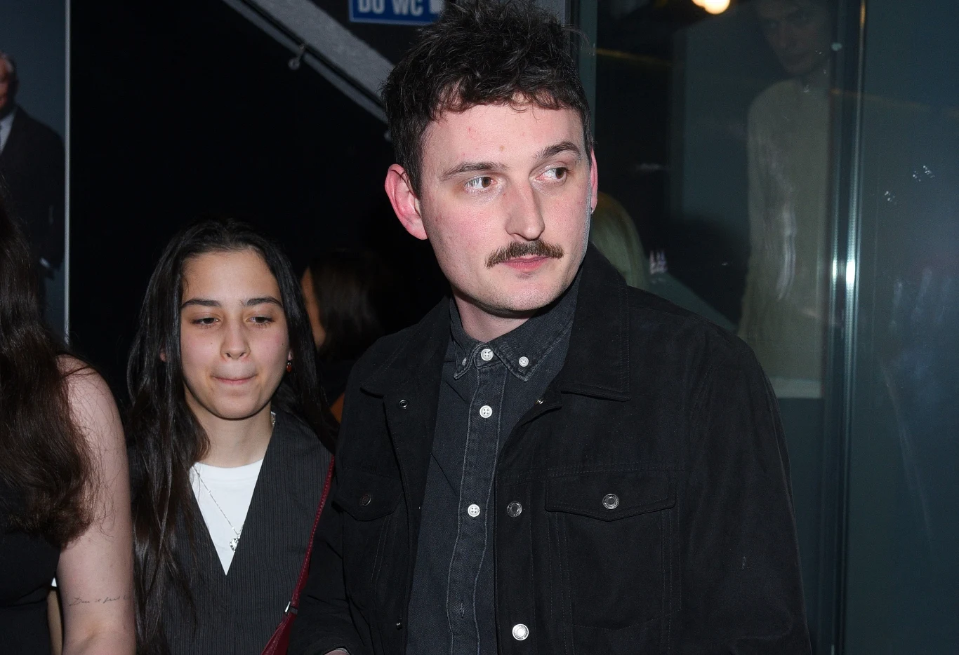 Dawid Podsiadło niespodziewanie pojawił się na koncercie The Fray w Warszawie Mężczyzna o ciemnych włosach i wąsach stoi obok młodej kobiety w jasnej bluzce i ciemnej marynarce, oboje znajdują się w pomieszczeniu ze szklaną ścianą i osobami w tle.