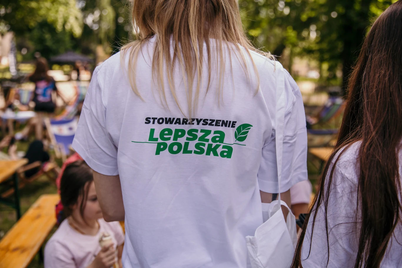 Czyste, Zielone Miasta w Lesznie Osoba w białej koszulce z napisem Stowarzyszenie Lepsza Polska stoi tyłem, wokół niej znajdują się inne osoby oraz drewniane ławki i stoły w parku, w tle widać zielone drzewa.