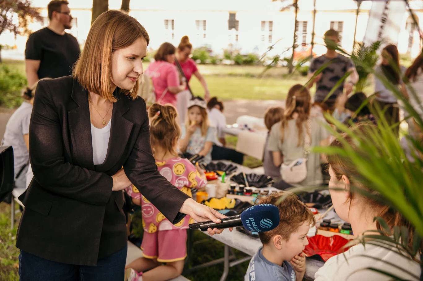 Czyste, Zielone Miasta w Lesznie Reporterka przeprowadza wywiad z kobietą i chłopcem podczas plenerowego wydarzenia dla dzieci, w tle widoczne stoły z przyborami plastycznymi oraz bawiące się dzieci.