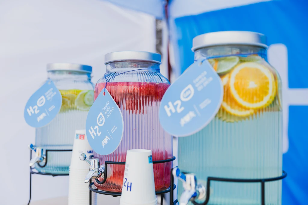 Czyste, Zielone Miasta w Gdańsku Trzy duże szklane słoje z wodą, w których znajdują się plasterki cytryny, pomarańczy, limonki i arbuza, stoją na metalowych stojakach, każdy z nich oznaczony niebieską etykietą z napisem H2O, obok ustawione są papierowe kubki.