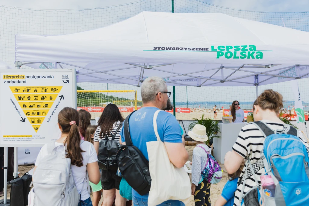 Czyste, Zielone Miasta w Gdańsku Grupa ludzi z plecakami i torbami stoi przed białym namiotem z napisem „Stowarzyszenie Lepsza Polska”. Obok widoczna tablica z hierarchią postępowania z odpadami. W tle siatka i fragment plaży.