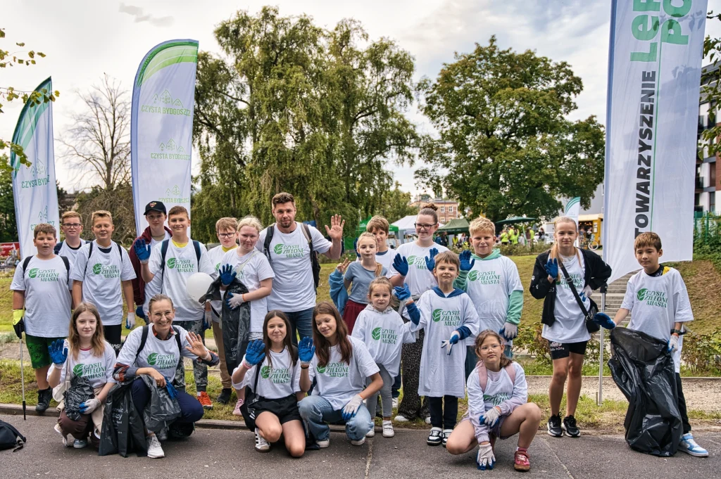 Czyste, Zielone Miasta w Bydgoszczy Grupa dzieci i młodzieży w białych koszulkach z logiem organizacji ekologicznej pozuje z workami na śmieci i rękawicami ochronnymi, uczestnicząc we wspólnej akcji sprzątania na świeżym powietrzu, na tle drzew i flag stowarzyszenia.