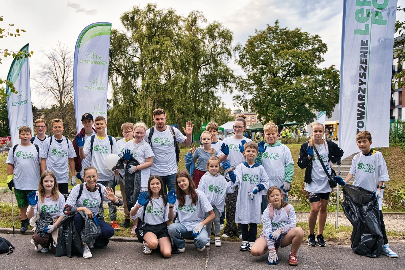 Czyste, Zielone Miasta w Bydgoszczy Grupa dzieci i młodzieży w białych koszulkach z logiem organizacji ekologicznej pozuje z workami na śmieci i rękawicami ochronnymi, uczestnicząc we wspólnej akcji sprzątania na świeżym powietrzu, na tle drzew i flag stowarzyszenia.