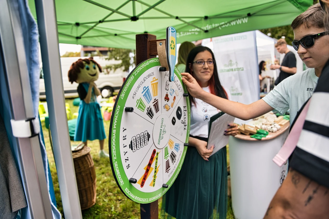 Czyste, Zielone Miasta w Bydgoszczy Stoisko edukacyjne z kołem fortuny dotyczącym segregacji odpadów, otoczone przez osoby biorące udział w zabawie. W tle namiot i maskotka ubrana w niebieską spódnicę.