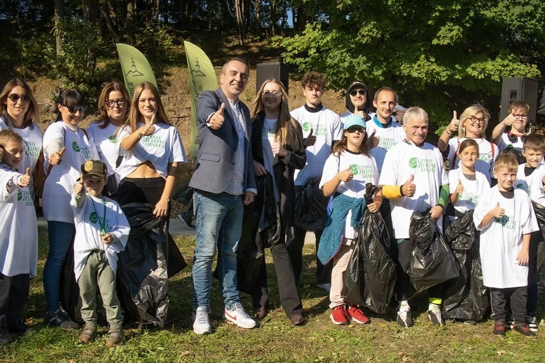 Czyste, Zielone Miasta w Zielonej Górze Grupa osób w różnym wieku ubrana w białe koszulki z napisem, stojąca razem na tle zieleni, wiele z nich trzyma czarne worki na śmieci i pokazuje kciuk do góry, atmosfera współpracy i zaangażowania.