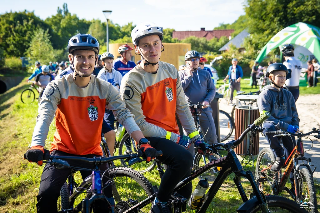 Czyste, Zielone Miasta w Zielonej Górze Dwóch młodych mężczyzn w pomarańczowych koszulkach i kaskach rowerowych siedzi na rowerach, uśmiechając się do obiektywu. Za nimi widoczne są inne osoby na rowerach, wskazujące na udział w wydarzeniu rowerowym lub zawodach w plenerze. W tle znajdują si...