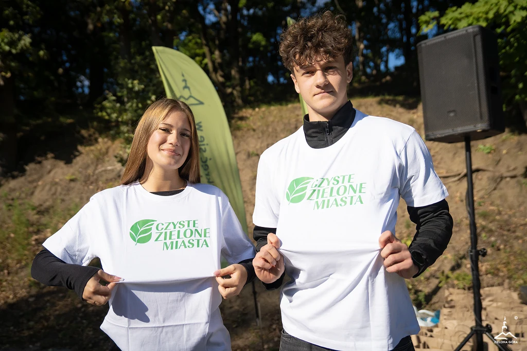 Czyste, Zielone Miasta w Zielonej Górze Dwie młode osoby w białych koszulkach z napisem 'Czyste Zielone Miasta' stoją na tle zieleni i trzymają rękoma materiał koszulek wyeksponowany do przodu, promując akcję ekologiczną w słoneczny dzień.