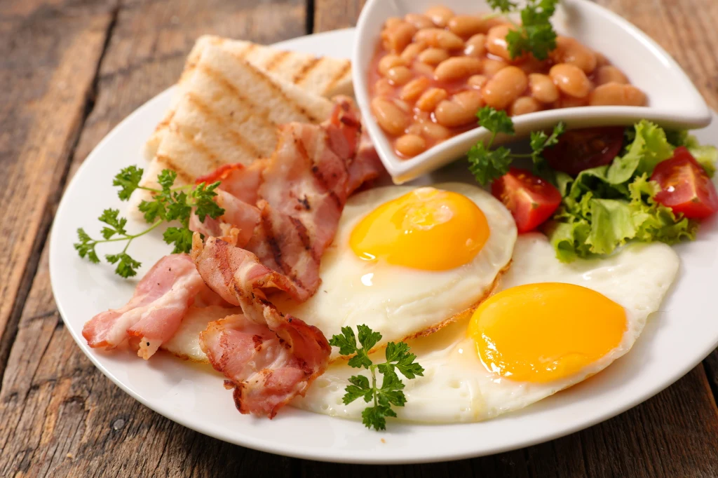 Pełne śniadanie z jajkami sadzonymi, grillowanym boczkiem, tostami, fasolką w sosie pomidorowym, sałatą oraz natką pietruszki na białym talerzu umieszczonym na drewnianym stole.