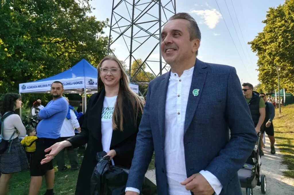 Czyste, Zielone Miasta w Zielonej Górze Mężczyzna i kobieta uśmiechają się, spacerując wśród ludzi na wydarzeniu plenerowym, w tle widoczne są drzewa, namioty oraz wysoki słup energetyczny.