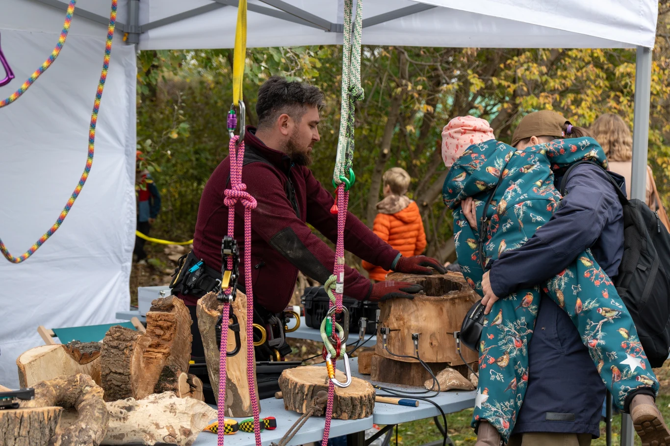 Czyste, Zielone Miasta w Warszawie Mężczyzna prowadzi warsztaty przy stole na świeżym powietrzu, obok stoi dorosła osoba trzymająca dziecko na rękach; na stole kawałki drewna, narzędzia i kolorowe liny, w tle widać inne osoby uczestniczące w wydarzeniu.