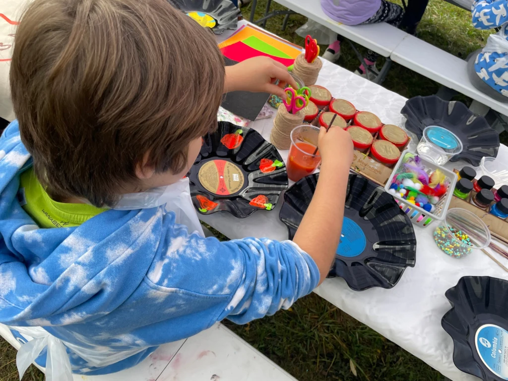 Czyste, Zielone Miasta w Warszawie Dziecko siedzi przy stole, wykonując prace plastyczne z użyciem różnych materiałów, takich jak kolorowe pompony, farby i dekoracyjne patyczki. Na stole stoją słoiczki z czerwoną przykrywką, pojemnik z koralikami oraz czarny talerz z rozłożonymi przybor...
