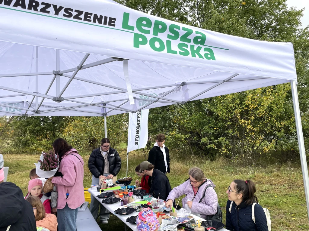 Czyste, Zielone Miasta w Warszawie Ludzie siedzą i stoją przy stolikach pod namiotem z napisem 'Lepsza Polska', zajmując się tworzeniem prac plastycznych lub rękodziełem na tle zielonej przestrzeni i drzew.