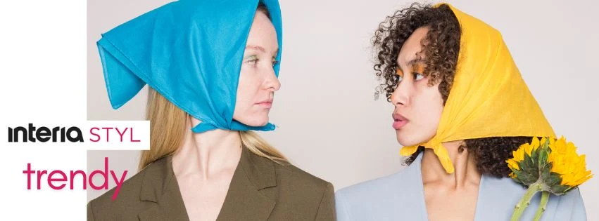 Dwie młode kobiety patrzą na siebie z powagą, jedna ubrana w niebieską chustę, druga w żółtą chustę, ubrana w jasną marynarkę i trzymająca słonecznik, w tle logo Interia Styl oraz napis trendy.