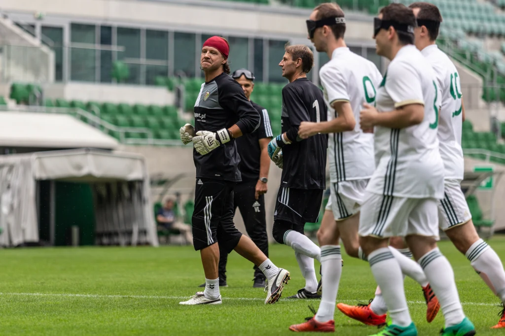 Drużyna piłkarska w białych strojach i opaskach na oczach maszeruje po murawie stadionu, z przodu widoczny bramkarz w rękawicach i czerwonej opasce na głowie. W tle trybuny stadionu.