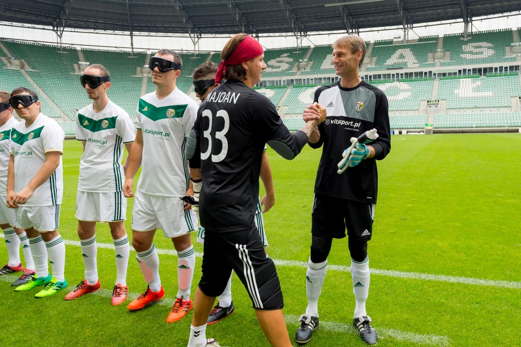 Piłkarze w białych strojach oraz bramkarze uściślający sobie dłonie na płycie stadionu piłkarskiego, część zawodników ma na oczach opaski, co wskazuje na mecz piłki nożnej dla osób niewidomych.