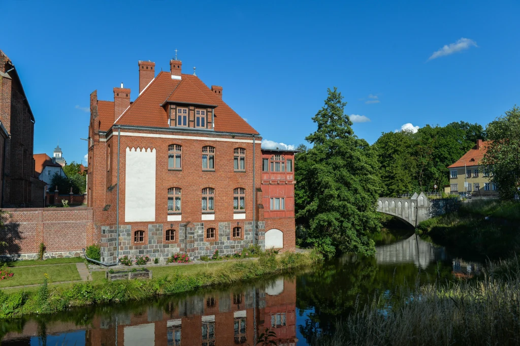 Przecinająca ulice miasta Łyna i zabytkowa architektura sprawiają, że Lidzbark zyskał miano "polskiej Wenecji" Ceglany budynek z czerwonym dachem położony nad rzeką, w tle kamienny most i zieleń drzew pod błękitnym niebem.