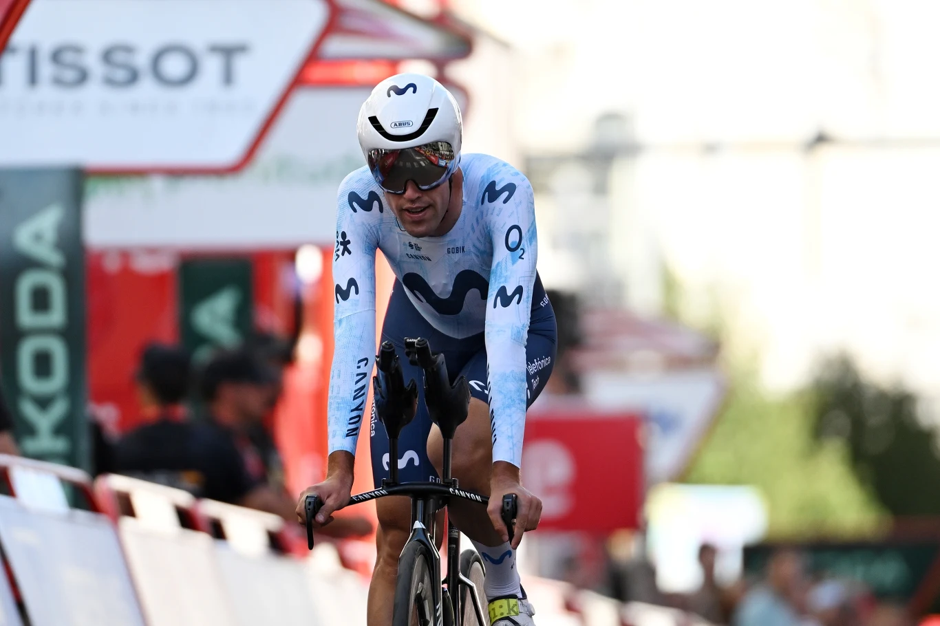 Kolarz w stroju wyścigowym jadący na rowerze podczas zawodów, z widocznym logotypem zespołu na stroju i kasku, w tle rozmyta publiczność i barierki oraz elementy oznaczeń sponsorskich.