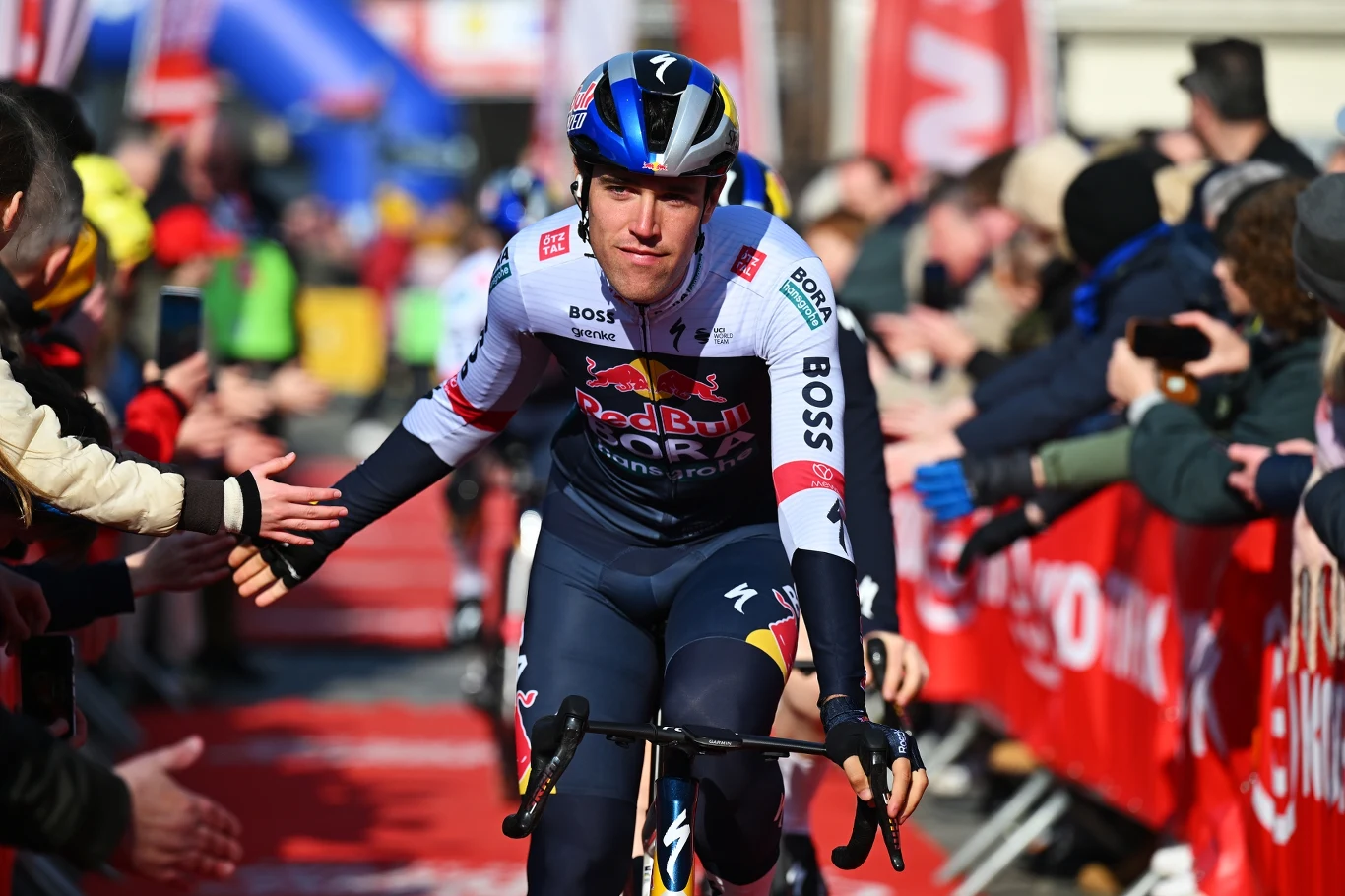 Zawodowy kolarz w stroju drużynowym Red Bull Boss na rowerze podczas wyścigu, jadący po czerwonym dywanie, przybijający piątki kibicom stojącym po obu stronach trasy, w tle widoczne tłumy i banery reklamowe.