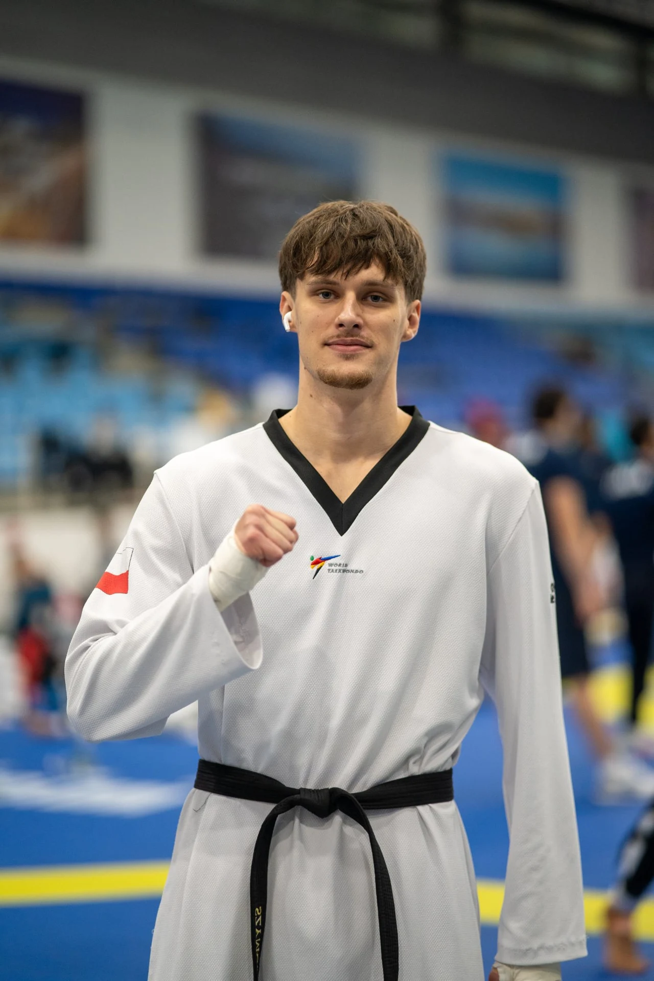 Młody mężczyzna w białym stroju do taekwondo z czarnym pasem, stoi na tle niebieskiej sali sportowej, wykonując gest pięści, w tle widoczne rozmyte postacie innych zawodników.